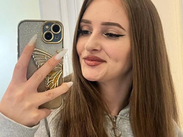 Pamela Xu, con lunghi capelli castani, indossa una felpa grigia, tiene in mano un telefono con una cover a forma di farfalla, sorride alla fotocamera, mostrando le sue unghie bianche e l'eyeliner ad ala di gabbiano..