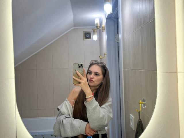 PamelaXu taking a mirror selfie, showing her tits in a grey hoodie, with long brown hair, and wearing eyeglasses on her head, in a brightly lit bathroom.