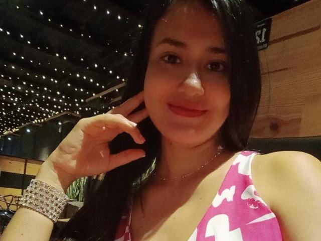 SarahMoon : Une femme aux cheveux foncés, au rouge à lèvres rouge et à la robe fleurie rose, touchant sa joue, portant un bracelet scintillant, souriant doucement. Ses seins sont visibles, sa main est près de son visage..