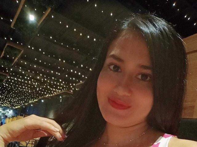 SarahMoonOne with dark, straight hair, red lips, and subtle makeup, smiling softly, hand near face, wearing a pink-and-white top, under string lights.