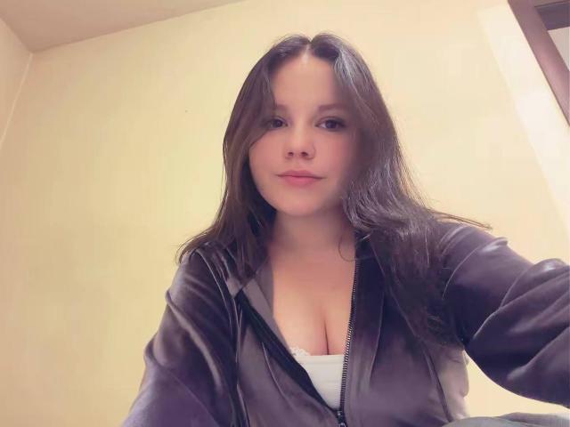 IolaLandauer has long brown hair, a slightly chubby face, and is wearing a black jacket with a white shirt showing her big, round tits. She's looking at the camera.