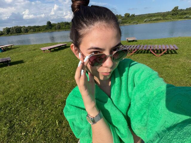 EmeraldCrivaro with a green towel, brown bun, and aviator sunglasses, holding her phone, showing off her white manicured nails and green eyes, wearing a green watch.