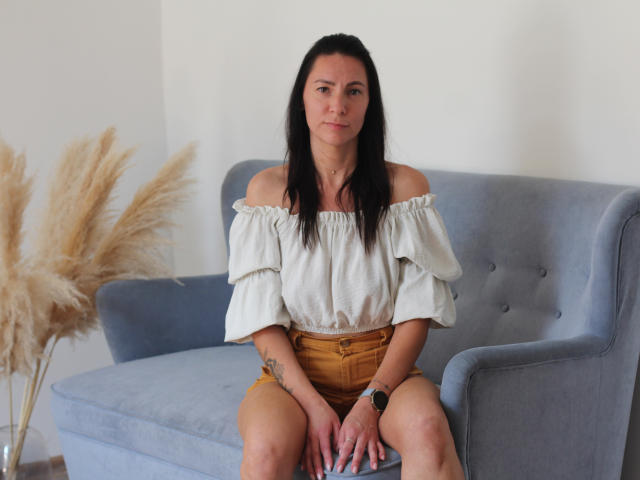IngaDrein, avec ses cheveux noirs, portant un haut blanc à épaules dénudées, un short jaune, assise sur un canapé bleu, avec de l'herbe tachetée de beige à gauche, portant une montre noire, avec un tatouage sur l'avant-bras droit..