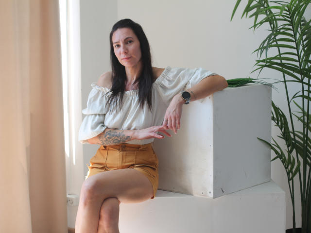 IngaDrein met lang zwart haar, een witte top met open schouder, en gele shorts, zittend op een grijze kubus, met een tatoeage op de linkeronderarm, een zwart horloge om, en een serieuze blik..
