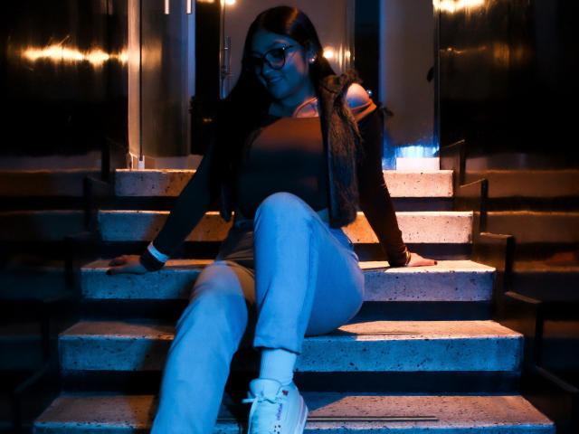 MiaaVixen with glasses, dark jacket, and blue jeans, sitting on concrete steps, legs crossed, white sneakers, black hair, blue light, arm resting on step, looking down.