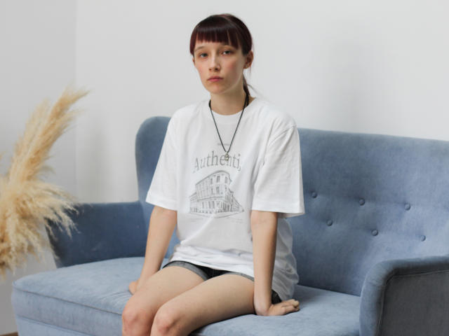 RiaVein, avec ses courts cheveux roux, son t-shirt blanc, son collier et son short noir, est assise sur un canapé bleu, avec une expression sérieuse. Sa queue, volumineuse, est visible en arrière-plan, sur la gauche. Les fesses de RiaVein sont partiellement visibles..