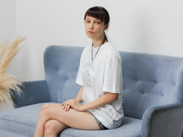 RiaVein, sentada em um sofá azul, vestindo uma camiseta branca e shorts cinzas, com um colar, tem uma expressão séria, e seu cabelo castanho está preso em um rabo de cavalo..