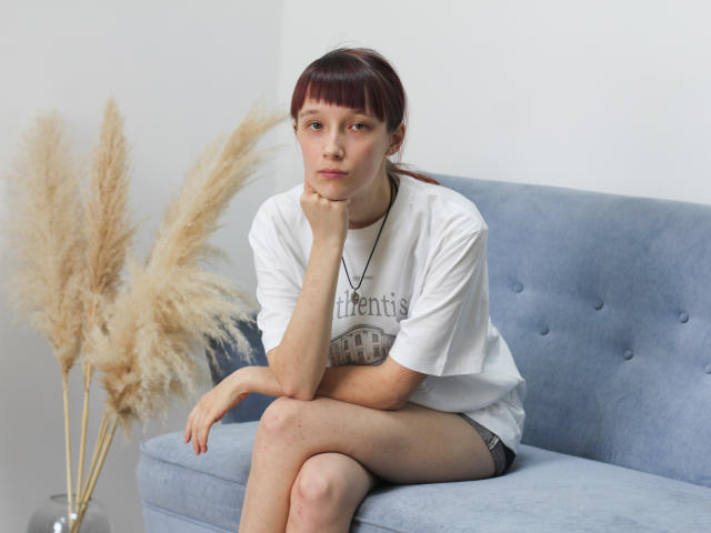 RiaVein, avec ses courts cheveux roux, son t-shirt blanc, son short noir et son collier, assise sur un canapé bleu, la main sur le menton, avec des herbes brunes et gonflées en arrière-plan, a l'air sérieuse, et a la peau pâle..