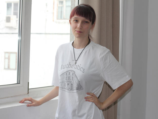 RiaVein met rode lokken, een wit T-shirt en een ketting, staat bij een raam, met haar handen op haar heupen, licht glimlachend, vooruitkijkend, natuurlijk licht, witte muren, beige gordijn..