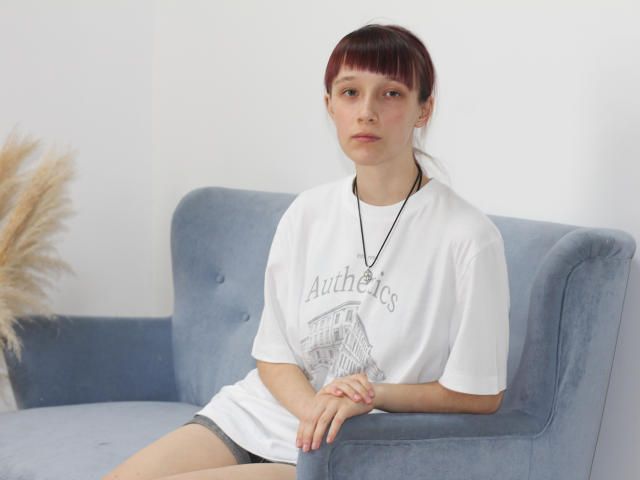 RiaVein, avec des franges brunes, portant un t-shirt blanc et un short gris, est assise sur un canapé bleu, les mains posées sur ses genoux, un collier, regardant droit devant elle, avec des ongles roses et gonflés..