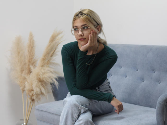 DataSley with blonde hair, glasses, green long-sleeve top, gray pants, and hand on cheek, sitting on blue sofa with pampas grass, looking thoughtful.