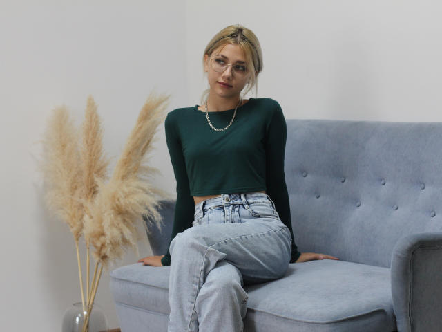 DataSley, con cabello rubio, gafas, un top corto verde y jeans azul claro, sentada en un sofá gris, con hierba de pampa color beige esponjosa en un jarrón de vidrio a su izquierda..
