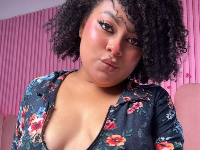 ElizabethBloom has curly black hair, dark skin, and is wearing a floral top with a low neckline, showing her boobs. She's looking at the camera with a pouty expression.