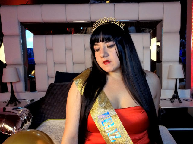SaraFountain with long black hair, red dress, gold sash, and crown, sitting on a bed, looking down, showing her tits, with a balloon in the foreground.