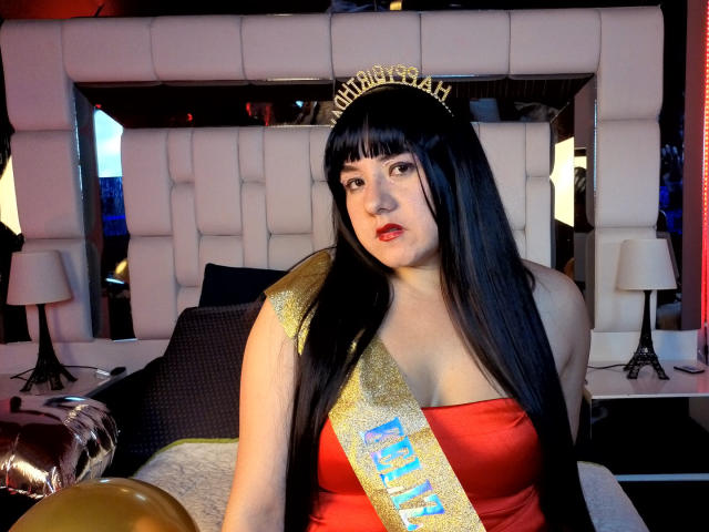 Sara, con lunghi capelli neri, vestito rosso, fascia dorata, corona e pelle chiara, seduta sul letto, che mostra il seno, con un palloncino dorato in primo piano..
