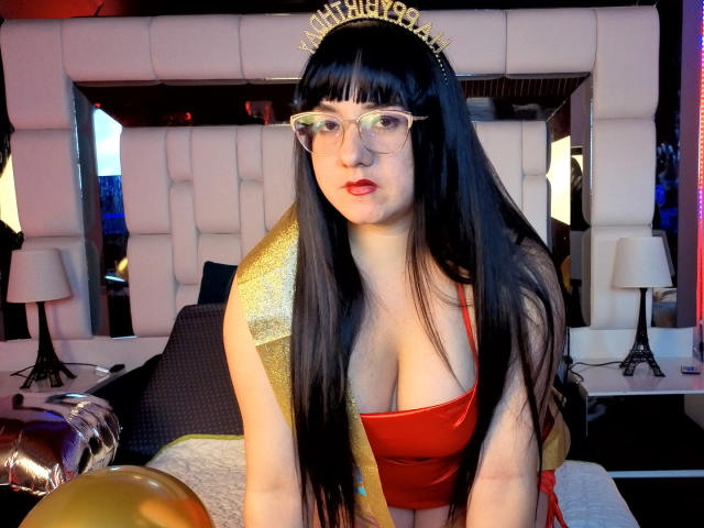 Sara Fountain mit langem, schwarzem Haar, Brille, rotem Kleid, goldener Haarreif und goldener Schulterdecke, die ihre großen Brüste zeigt, in einem Raum mit Lampen und Ballons..