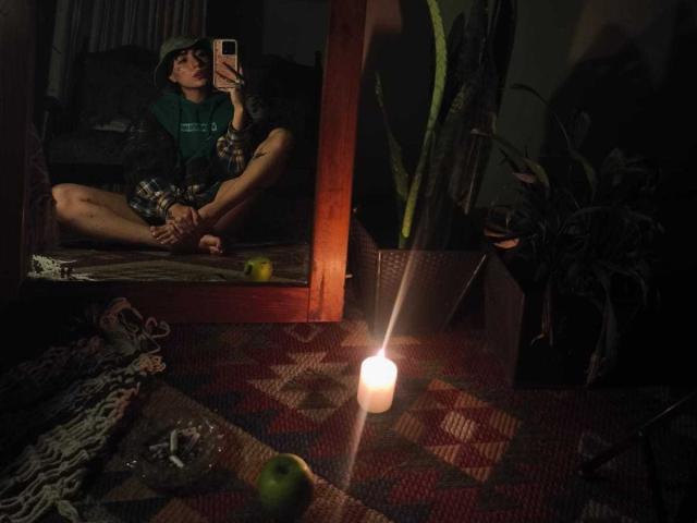 CleoZapata est assise par terre, les jambes croisées, devant un miroir, et elle prend un selfie avec son téléphone. Elle porte une chemise verte et un short à carreaux, et une bougie est allumée sur le sol..