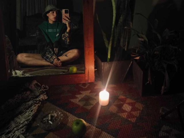 CleoZapata, assise en tailleur, se reflète dans un miroir, portant une chemise et un chapeau verts, dans une pièce éclairée par des bougies, avec une pomme verte et une plante en pot, et montrant un tatouage sur sa cuisse..