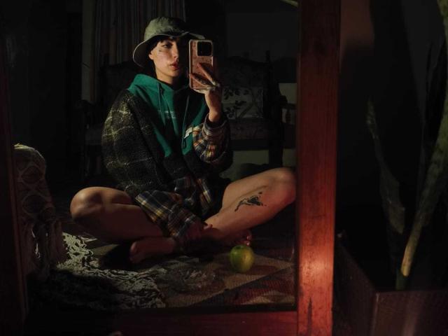 CleoZapata est assise par terre, les jambes croisées, devant un miroir, portant une écharpe verte, une chemise à carreaux et un chapeau de pêche, et elle prend un selfie avec un téléphone. Une pomme verte est sur le sol. Tatouage sur la cuisse..