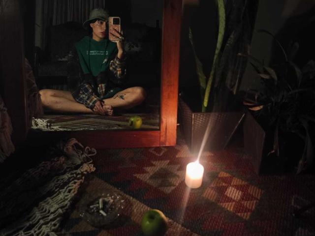 CleoZapata, assise en tailleur, portant une chemise verte avec des manches à carreaux, un chapeau, prend un selfie devant un miroir, montrant ses fesses nues, dans une pièce faiblement éclairée avec une bougie et une pomme verte..