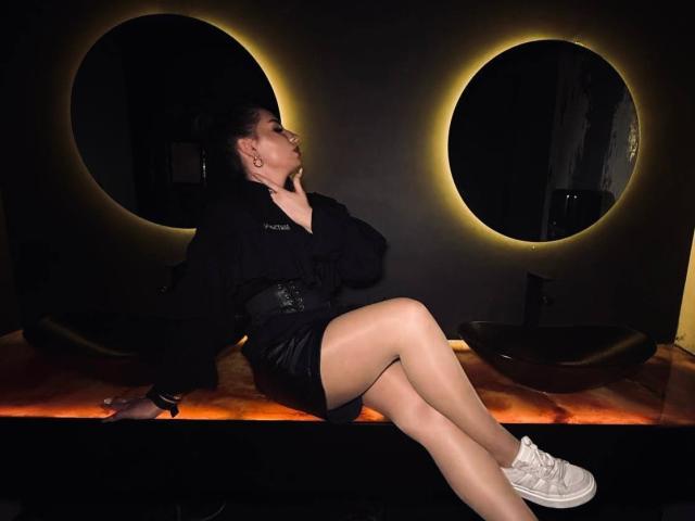 MarquitaVoltz with her tits slightly covered, sitting on a bench, wearing a black jacket, black shorts, sheer tights, and white sneakers, looking up, in a dark room.