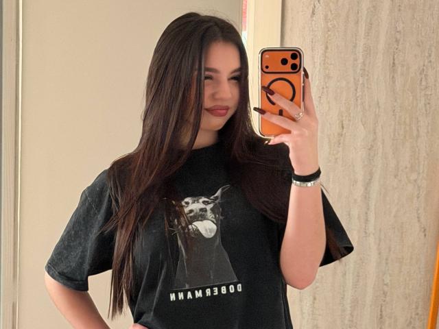 MarquitaVoltz with long brown hair, wearing a black Hammerhead t-shirt, holding an orange phone, taking a mirror selfie, smiling slightly, with a hand on her hip.