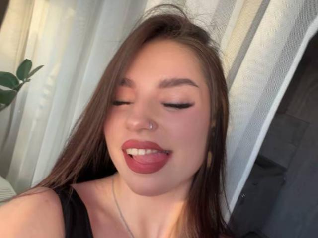 MarquitaVoltz with long brown hair, nose ring, and pink lipstick, eyes closed, tongue out, smiling, wearing black top, white curtains in background.