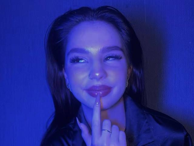 MarquitaVoltz with blue-lit face, dark wet hair, nose ring, and finger to lips, smiling slightly, wearing a black satin jacket, looking cute and mysterious.
