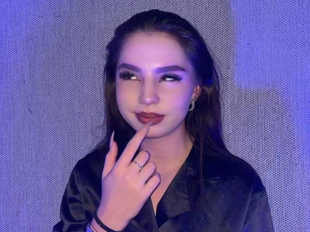 MarquitaVoltz with long brown hair, dark makeup, and purple eyeshadow, touching her lips with a finger, wearing a black satin shirt, and a ring. Blue lighting.