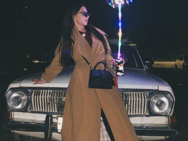 MarquitaVoltz stands in front of a white car, wearing a tan coat, black purse, holding a glowing stick, with long dark hair and sunglasses, looking cool.