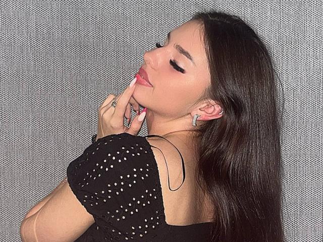 MarquitaVoltz with long dark hair, eyes closed, pink lipstick, black polka dot top, finger touching lips, ring on finger, earrings, gray textured background, sexy, thoughtful expression.