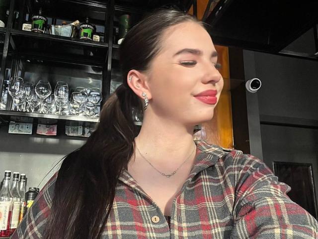 MarquitaVoltz has long brown hair in a ponytail, wearing a red plaid shirt, red lipstick, and a necklace, smiling with closed eyes, in a bar.