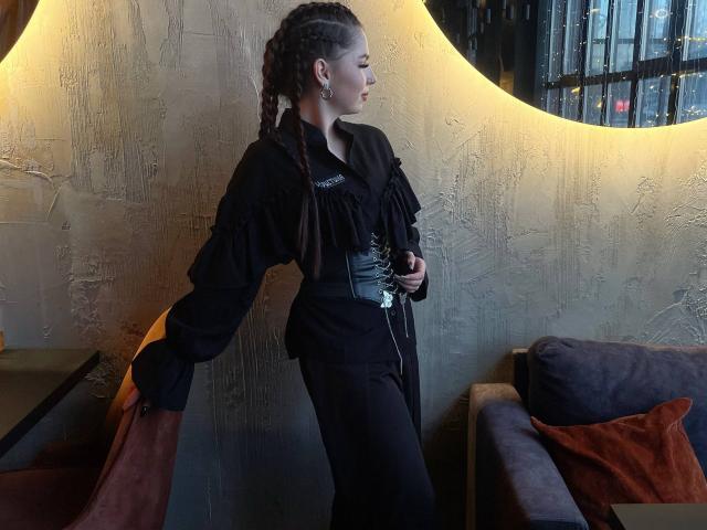 MarquitaVoltz with braided hair, black ruffled shirt, and black pants, standing by a curved, glowing wall, holding a phone, with a gray couch and orange pillow in background.