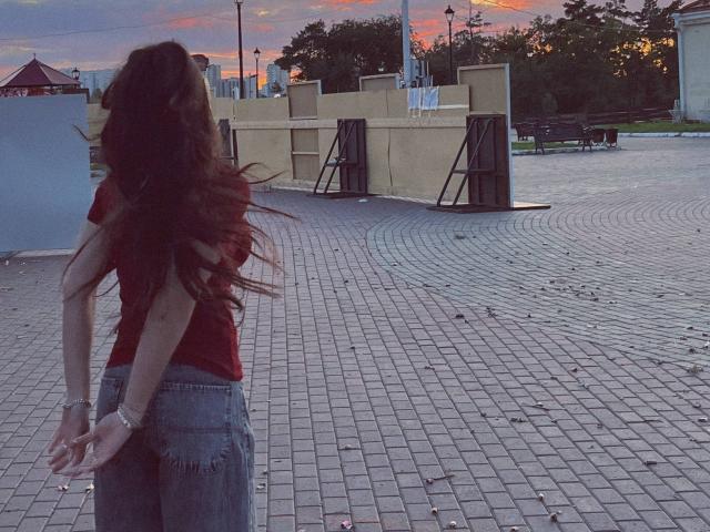 MarquitaVoltz with long brown hair in a red shirt and ripped jeans, standing with arms behind back, facing away, in a cobblestone plaza at sunset.