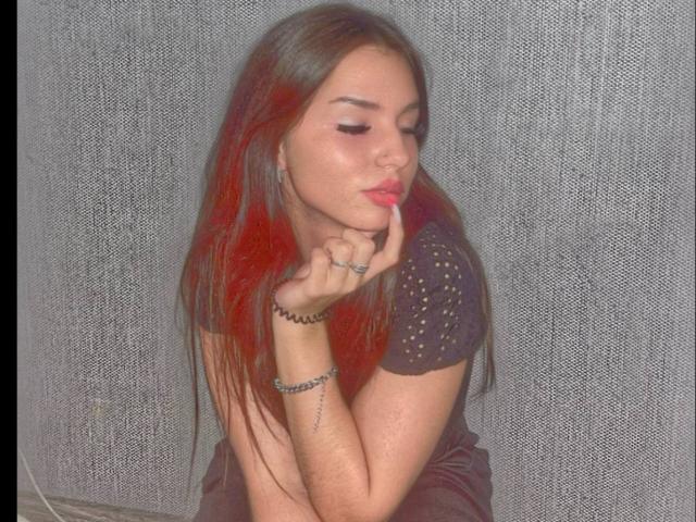 MarquitaVoltz with red-tinted brown hair, pouty lips, and finger to mouth, wearing a black, dotted top, sitting, showing off her ring and bracelet, looking sexy.