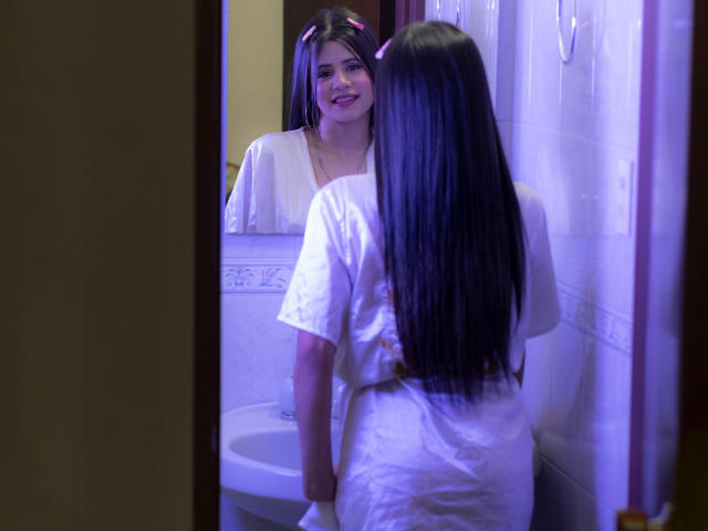 SheylaSpooner with long black hair, pink hair clips, smiling at herself in a mirror wearing a white shirt, reflected in a bathroom with purple lighting.