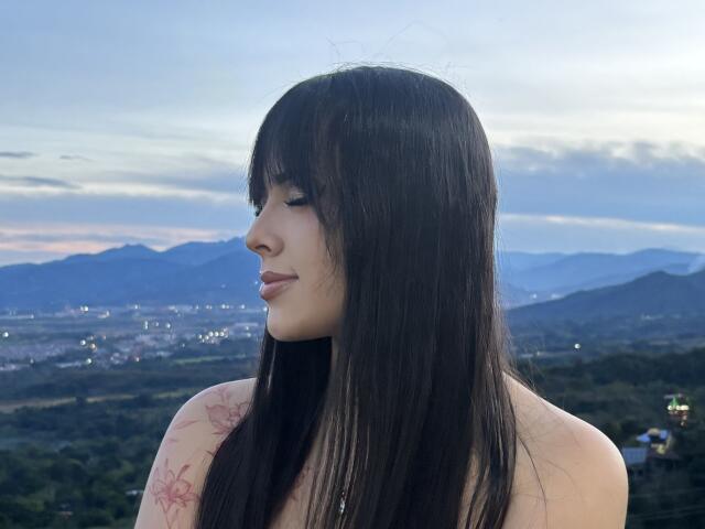 AlisonTatcher with long black hair, tattoo on left shoulder, pale skin, small nose, closed eyes, subtle smile, bare shoulders, soft evening light, mountains and city lights in background.