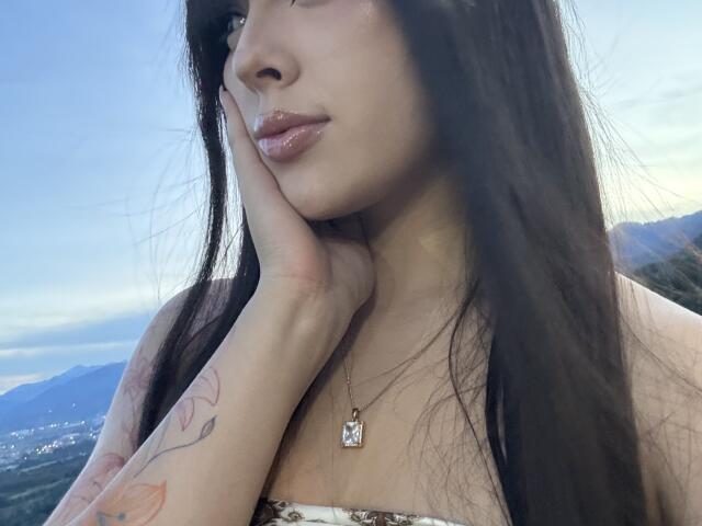 AlisonTatcher's face, with long brown hair, shiny pink lips, hand on cheek, tattoo on forearm, necklace with "M" pendant, wearing a strapless top, against a blue sky.