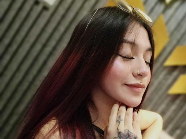 AngelsPhoenix has dark, red-tipped hair, closed eyes, and a subtle smile. She's wearing sunglasses on her head, has nose rings, and tattoos on her fingers.