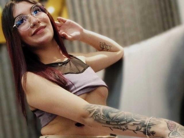 AngelsPhoenix with glasses, purple top exposing her small tits, tattooed arm, and red hair, smiling, hand near face. Her tattoos are on her left arm and right wrist.
