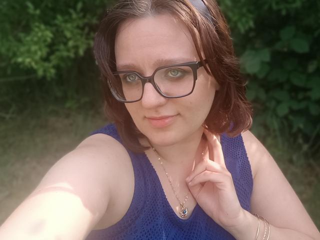 SensualParis mit braunen Haaren, Brille, blauem Top ohne Ärmel, Halskette, lächelnd, die Schlüsselbeine haltend, bei Tageslicht, wobei ihre Brüste im Bild leicht zu sehen sind, grünes Laub im Hintergrund..