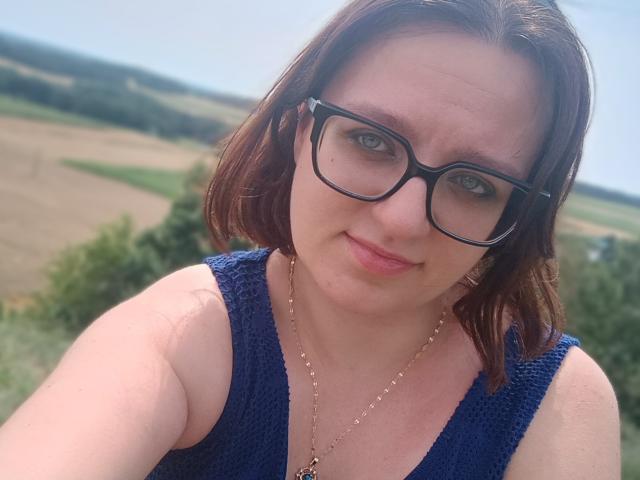 SensualParis, com cabelo castanho médio, óculos pretos, blusa de tricô azul e colar dourado, sorrindo suavemente, mostrando seios pequenos, em pé ao ar livre..