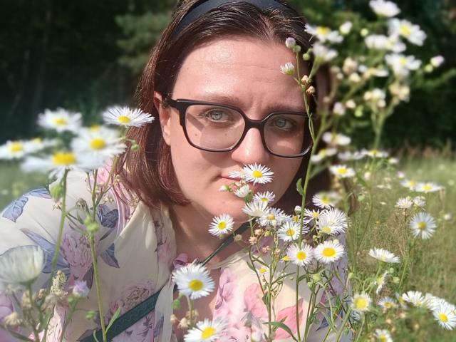 SensualParis met een bril, bruin haar en een bloes met bloemen, die een madelief houdt, staand in een veld met witte en gele bloemen, zonlicht op haar gezicht..