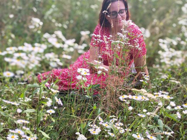 SensualParis, with glasses, pink floral dress, and brown hair, is lying in a field of white daisies, surrounded by green grass, looking down at the flowers.