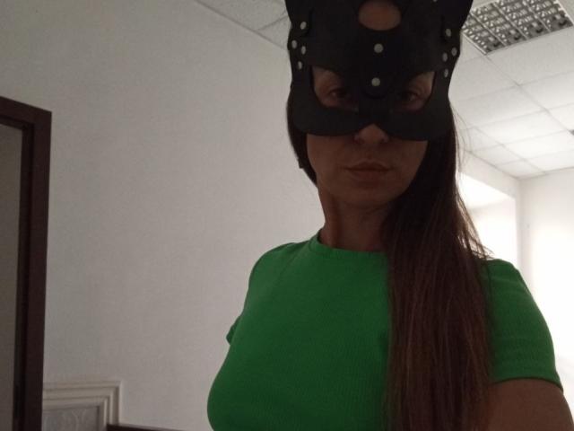 ViktoriiaLu wearing a black mask with eye holes, green shirt, and long brown hair, showing her tits slightly. She's looking down with a neutral expression.
