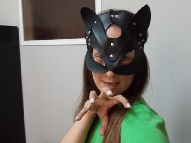 ViktoriiaLu wearing a black leather cat mask with metal studs, green shirt, long brown hair, white nails, and smiling at the camera with hands near her face.