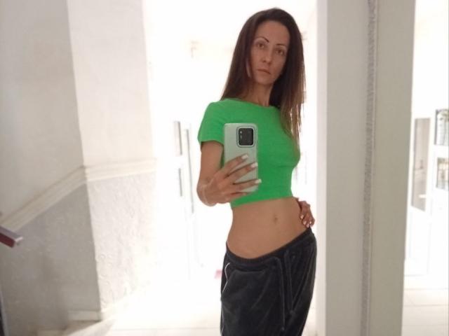 ViktoriiaLu with long brown hair, green crop top, showing her flat stomach, and black pants, taking a mirror selfie with her phone, in a bright room.
