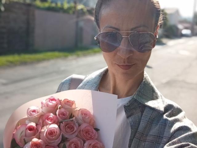 ViktoriiaLu with pink roses, wearing sunglasses, plaid shirt, and white tee, holding bouquet, street in background, sunlight, slight lens flare, subtle smile.