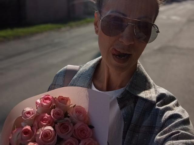 ViktoriiaLu with pink roses in a white bag, wearing a green plaid shirt and aviator sunglasses, smiling slightly. Sunlight casts a shadow on her face.