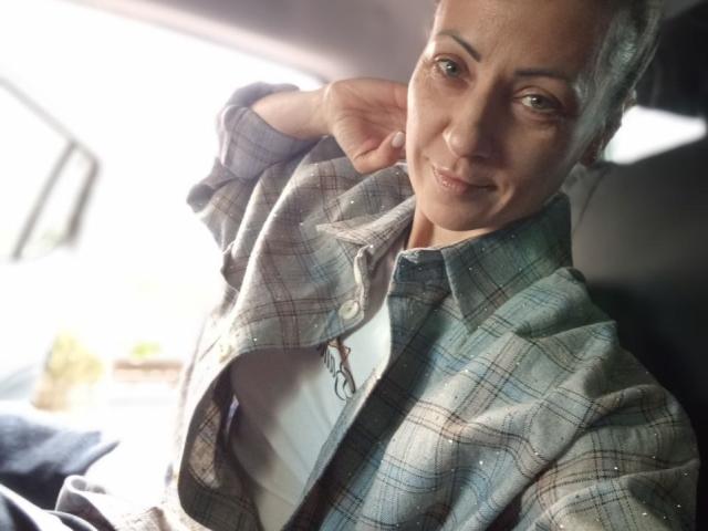 ViktoriiaLu, in a plaid shirt, leans back in a car, smiling with green eyes, and one hand behind her head. Her shirt is slightly open, showing a bit of chest.